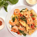 A white plate with Mediterranean Shrimp Pasta. A sprig of basil, some red pepper flakes and parmesan cheese surround the plate