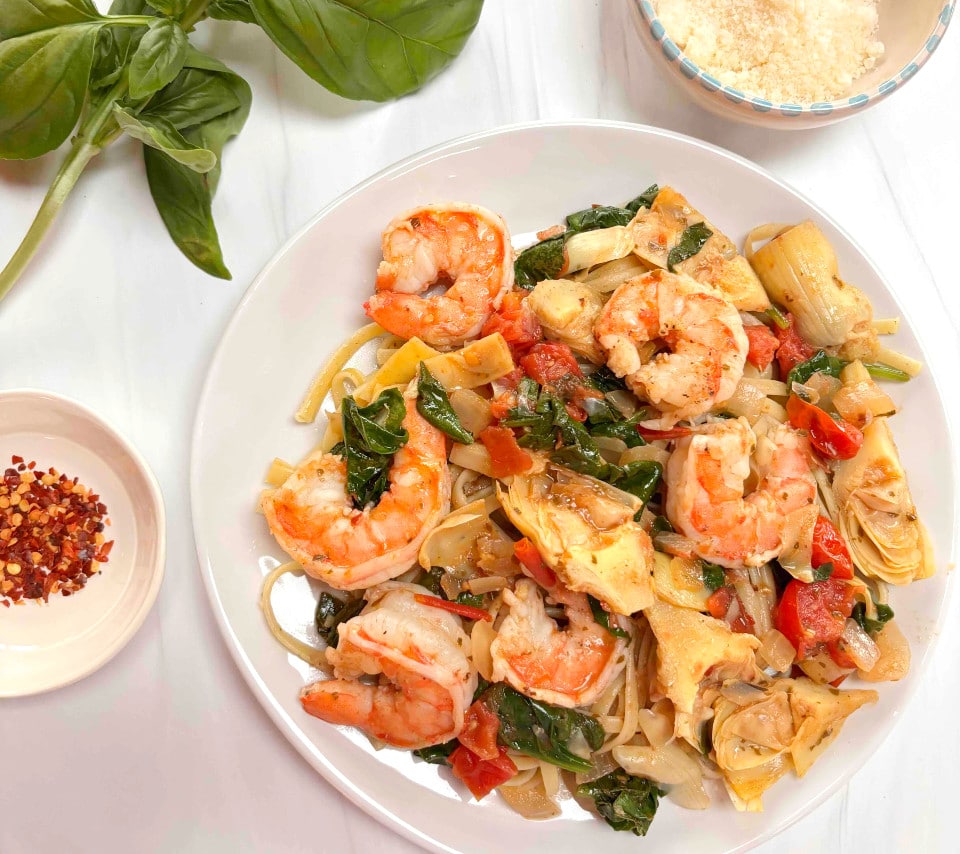 A white plate with Mediterranean Shrimp Pasta. A sprig of basil, some red pepper flakes and parmesan cheese surround the plate