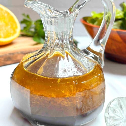 A small glass carafe filled with orange balsamic vinaigrette on a white counter. A cut orange and a salad are in the background.