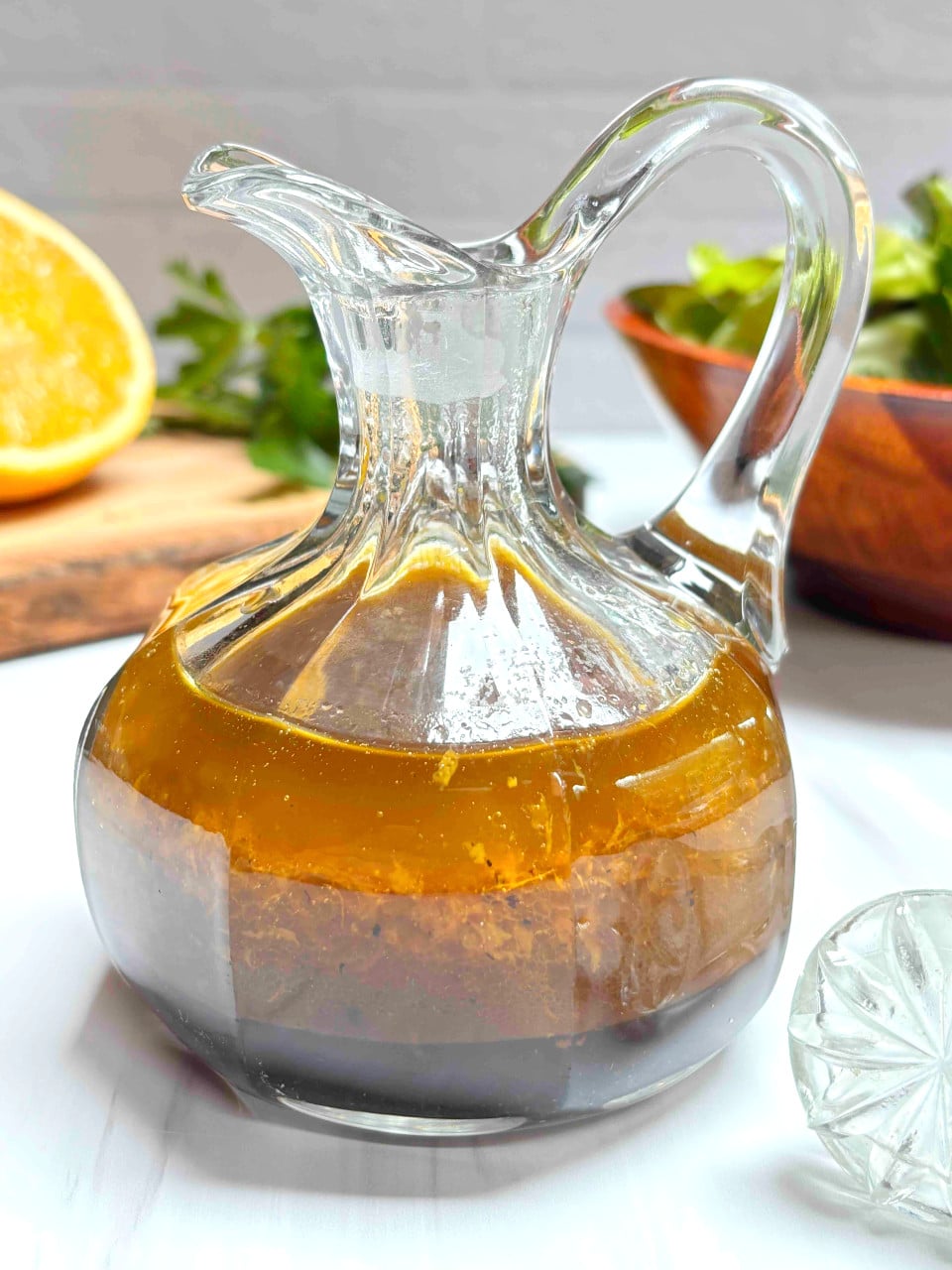 A small glass carafe filled with orange balsamic vinaigrette on a white counter. A cut orange and a salad are in the background.