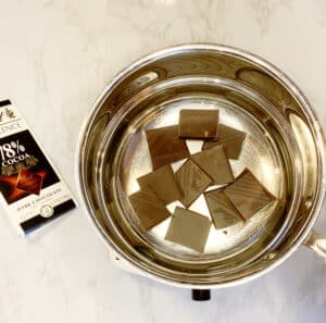 A metal pan with chopped dark chocolate in it, sitting on a burner. The pan is set on top of another pan with water. 