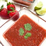 Two glass bowls of strawberry gazpacho soup on a white picnic table with strawberries and limes on the side.