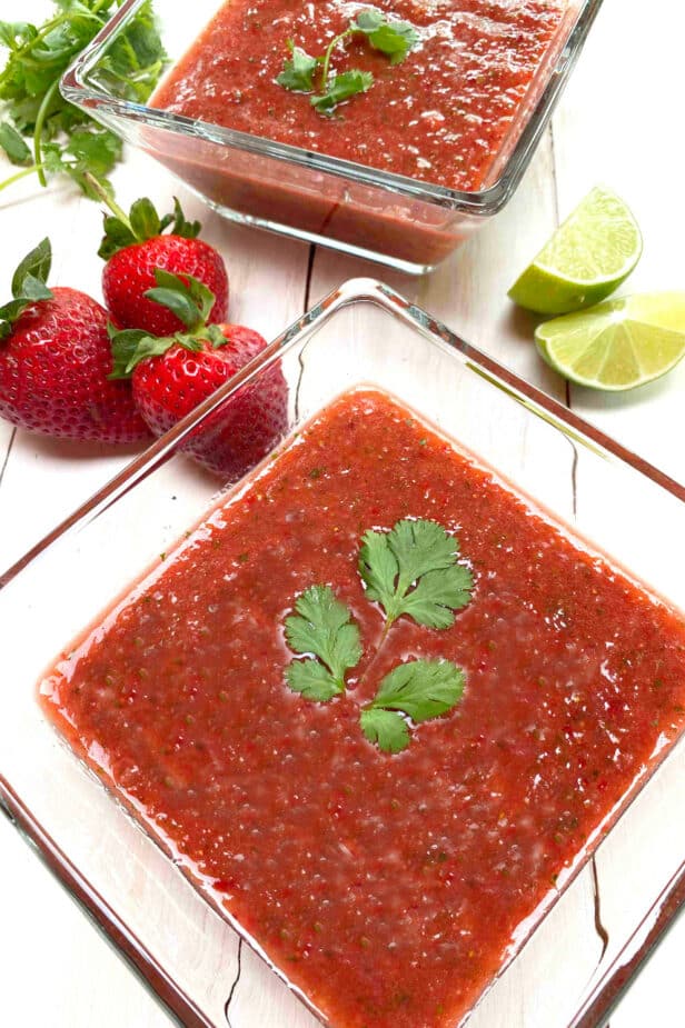 Two glass bowls of strawberry gazpacho soup on a white picnic table with strawberries and limes on the side.