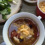 A white bowl filled with chicken tortilla soup on a wooden table with cilantro and garlic on a cutting board and shredded cheese in a bowl