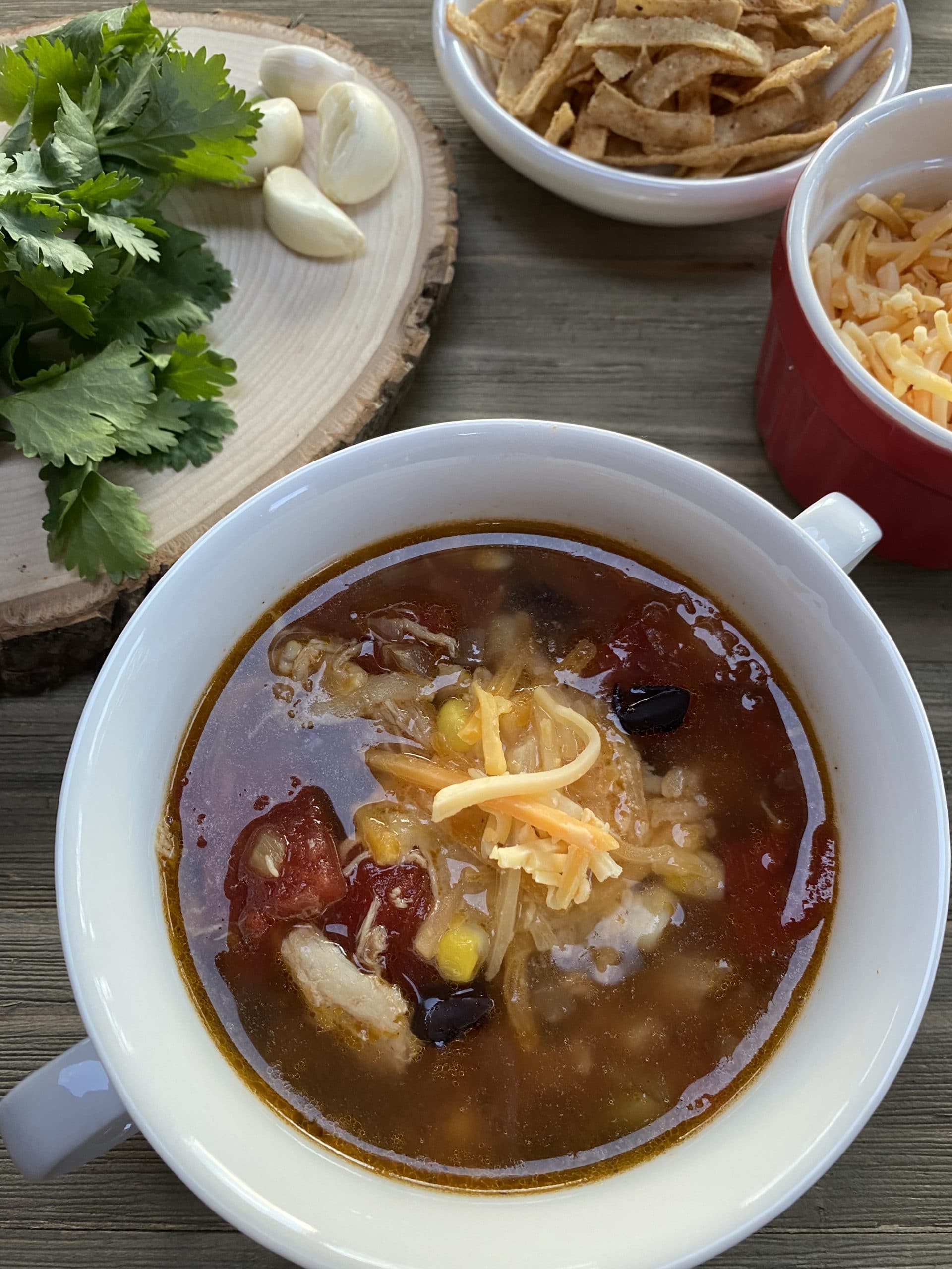 A white bowl filled with chicken tortilla soup on a wooden table with cilantro and garlic on a cutting board and shredded cheese in a bowl