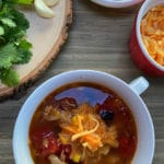 A white bowl filled with chicken tortilla soup on a wooden table with cilantro and garlic on a cutting board and shredded cheese in a bowl