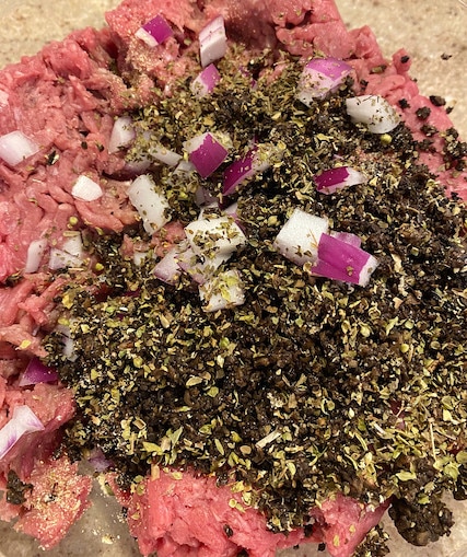 A bowl of raw ground beef topped with cooked, chopped mushrooms, red onions and Italian seasonings ready to be mixed into burgers.