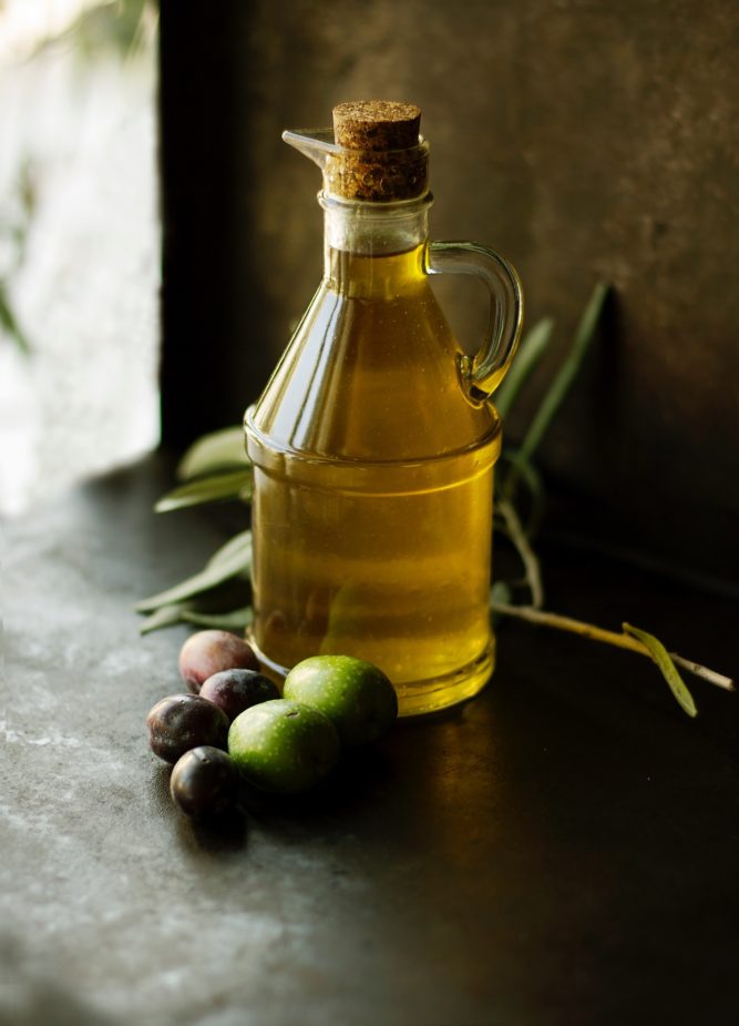 A carafe of olive oil on a dark background with olives and olive branches around it.
