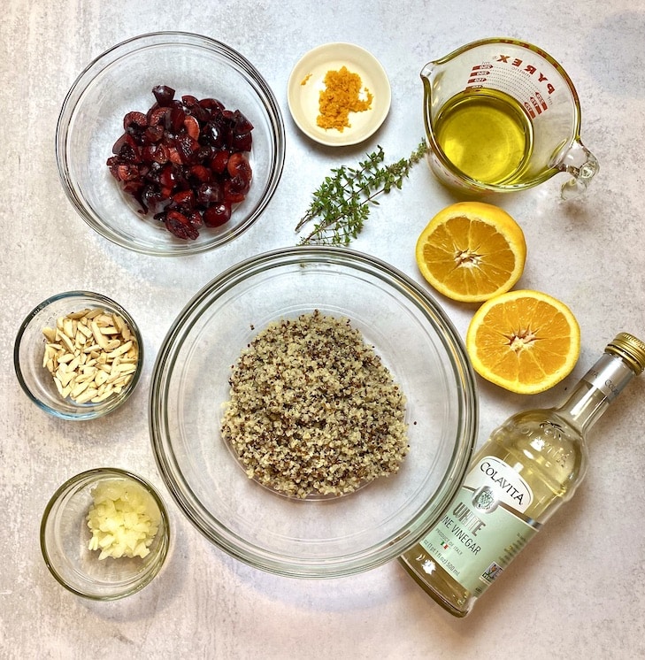 A counter with the ingredients used to make a quinoa salad including quinoa, almonds, cherries, oranges, vinegar and fresh thyme