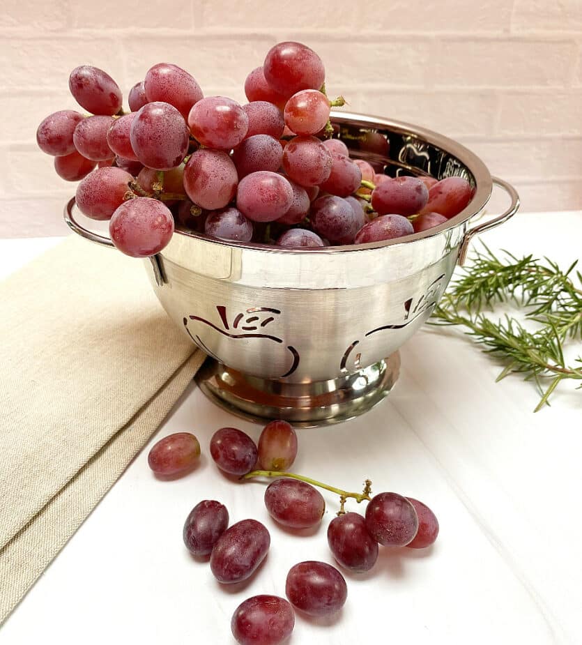 red grapes in a colandar on a white board with rosemary in the background