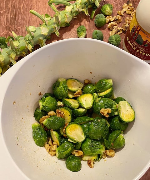 Halved Brussel sprouts in a white bowl tossed with oil, vinegar, maple syrup and walnuts