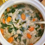 A white soup tureen filled with a chicken broth based soup with chicken, carrots, spinach and baby kale on a white table with cut lemons and bread in the background.