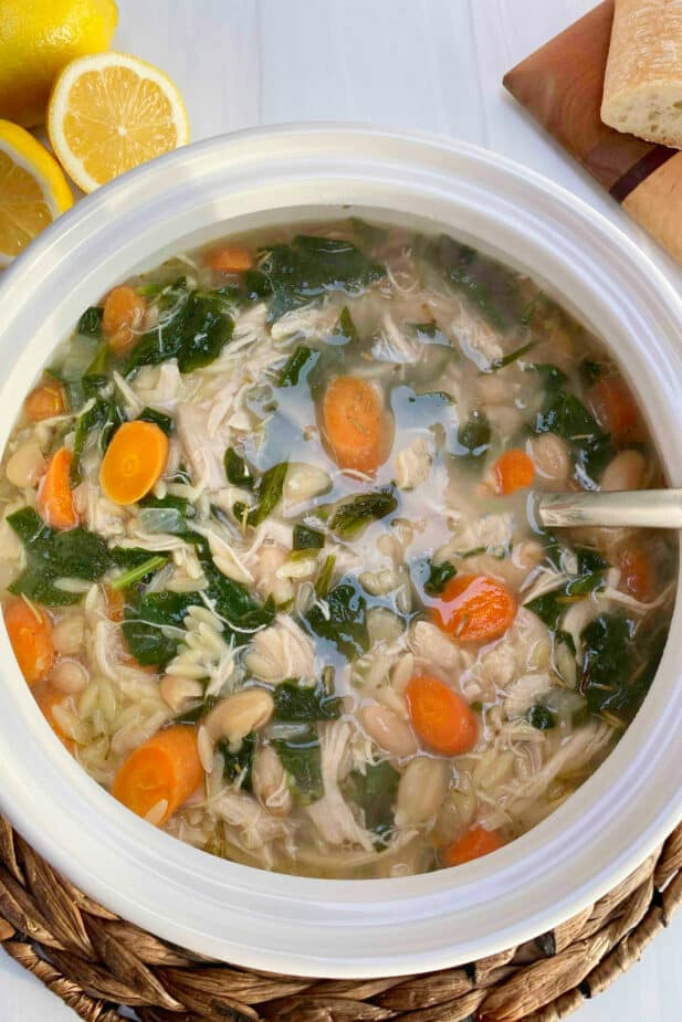 A white soup tureen filled with a chicken broth based soup with chicken, carrots, spinach and baby kale on a white table with cut lemons and bread in the background.