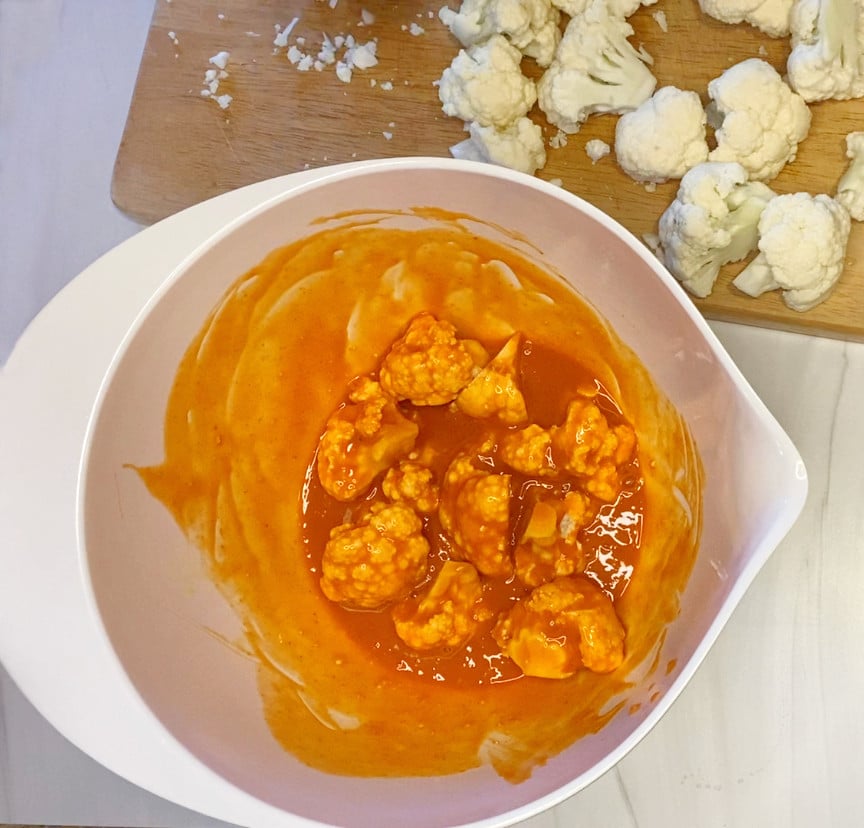 Cauliflower flowerets mixed in egg and hot Buffalo sauce in a white bowl.