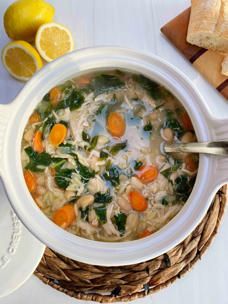 A white soup tureen filled with lemon chicken and orzo soup on a white counter with lemons and crusty bread on the side.