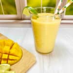 A yellow smoothie in a clear cup in front of a window with a cut mango and lime wedges on a wooden cutting board nearby.