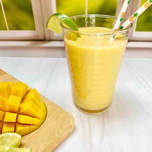 A yellow smoothie in a clear cup in front of a window with a cut mango and lime wedges on a wooden cutting board nearby.