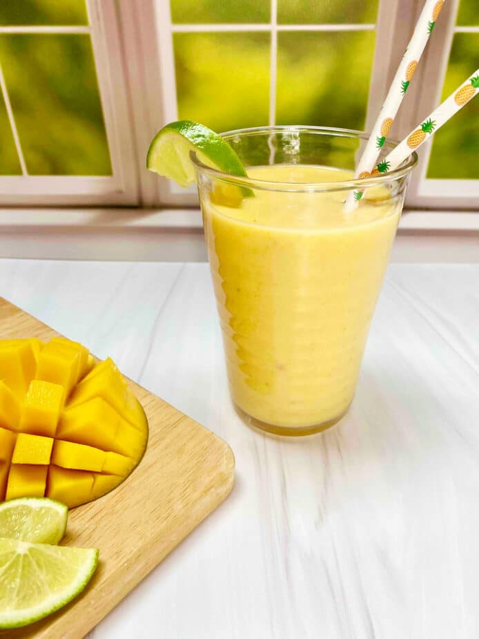 A yellow smoothie in a clear cup in front of a window with a cut mango and lime wedges on a wooden cutting board nearby.