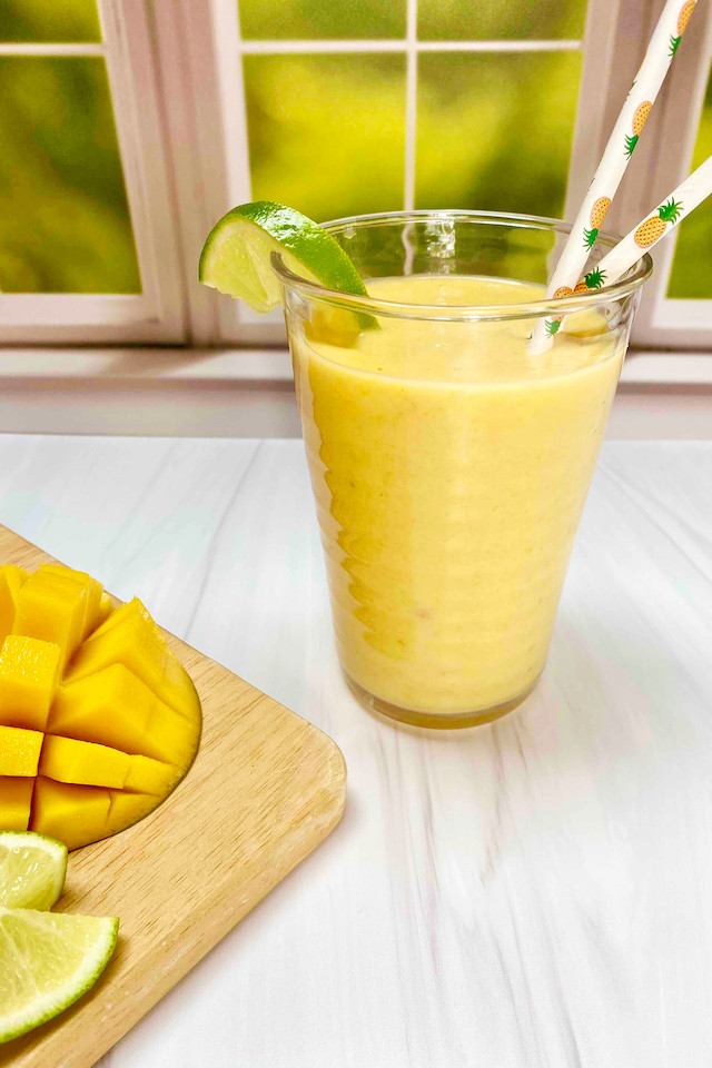 A yellow smoothie in a clear cup in front of a window with a cut mango and lime wedges on a wooden cutting board nearby.