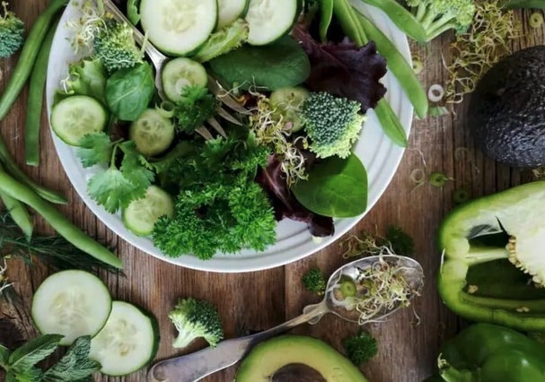 A white bowl filled with salad greens on a wooden cutting board surrounded by cut cucumbers and avocados