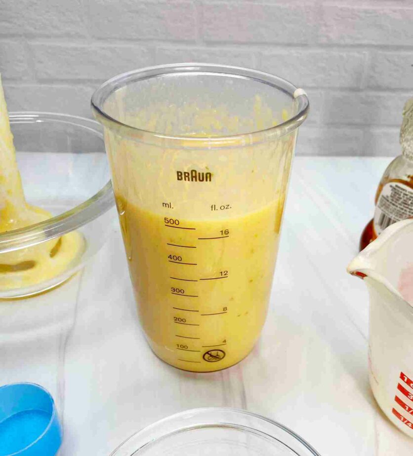 A blended mango kefir smoothie in a clear plastic cup on a white counter with an empty measuring cup and bowls in the foreground and blender stick in the back.