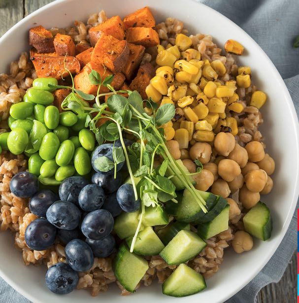 A white bowl filled with farro, blueberries, beans, corn avocado, edamame, and roasted sweet potatoes