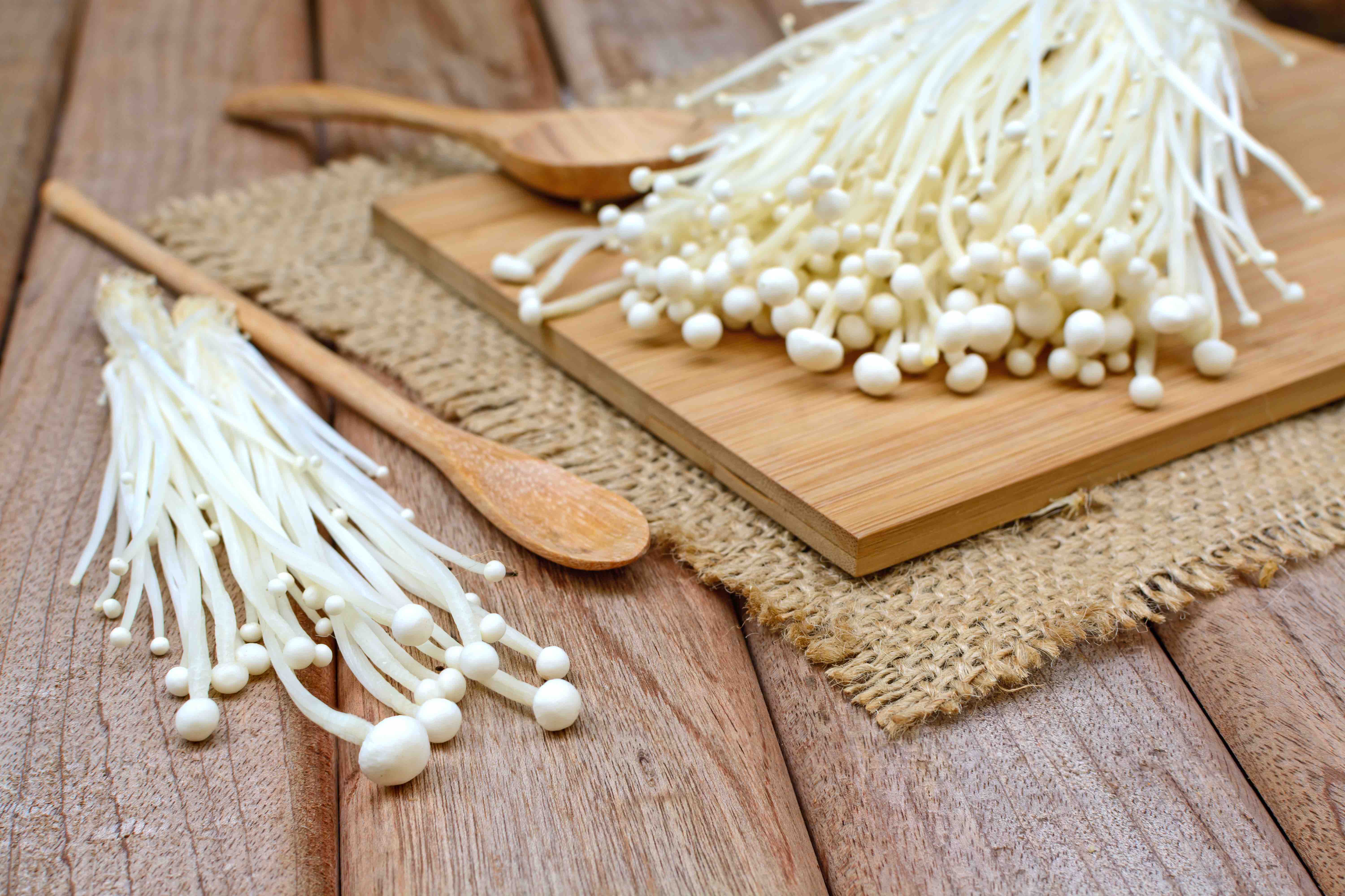 Enoki mushrooms on a wooden picnic table on a burlap mat and wood cutting board. A wooden spoon is on the side.