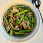 A white bowl of asparagus mushrooms and onion saute on a marble counter top with a serving spoon next to it.