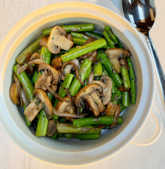 A white bowl of asparagus mushrooms and onion saute on a marble counter top with a serving spoon next to it.