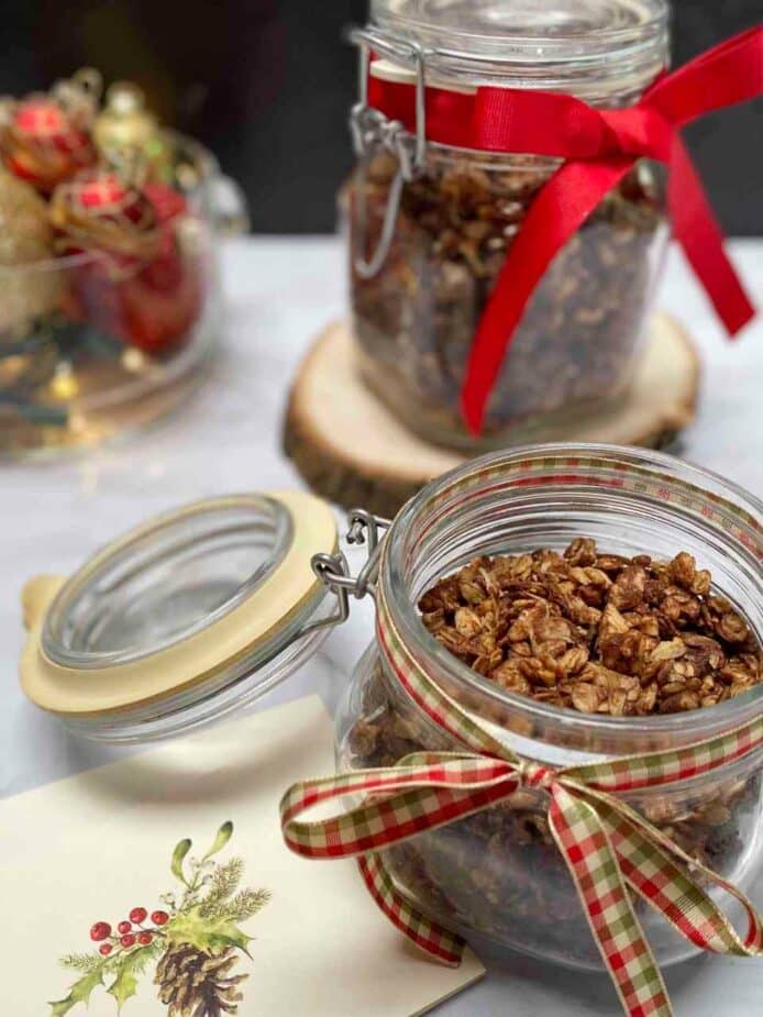 A small glass jar filled with Dark Chocolate Hazelnut Granola with a christmas ribbon around it. A Larger glass jar is behind it with a bowl of ornaments.