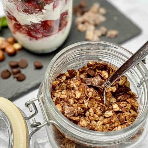 A glass jar filled with dark chocolate hazelnut granola with a jar of a yogurt parfait with plain yogurt and strawberries with the granola on a slate board.