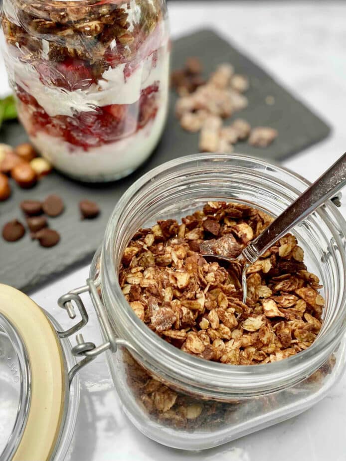 A glass jar filled with dark chocolate hazelnut granola with a jar of a yogurt parfait with plain yogurt and strawberries with the granola on a slate board.