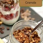 A glass jar filled with dark chocolate hazelnut granola with a jar of a yogurt parfait with plain yogurt and strawberries with the granola on a slate board.