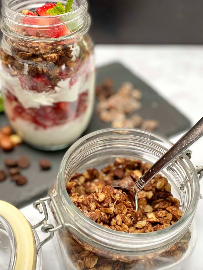 A glass jar filled with dark chocolate hazelnut granola with a jar of a yogurt parfait with plain yogurt and strawberries with the granola on a slate board.