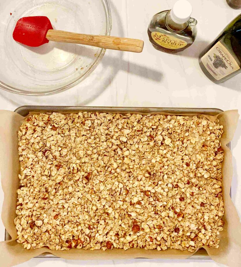 Granola spread on a baking sheet lined with parchment paper on a white marble counter top. An empty bowl and bottle of maple syrup and grapeseed oil are off to the side.