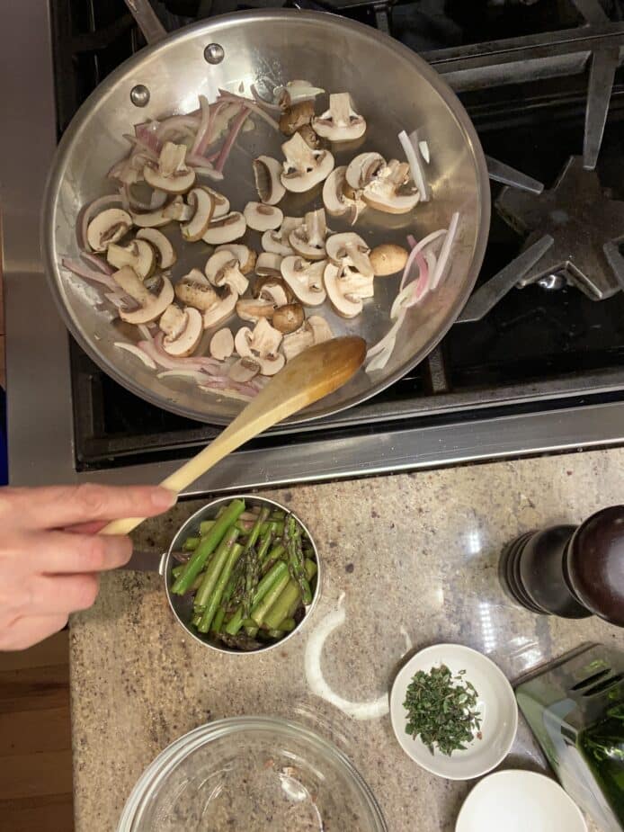 Mushrooms and onions cooking in a large sautepan on a stove. The other ingredients are sitting on the side and are ready to be added.