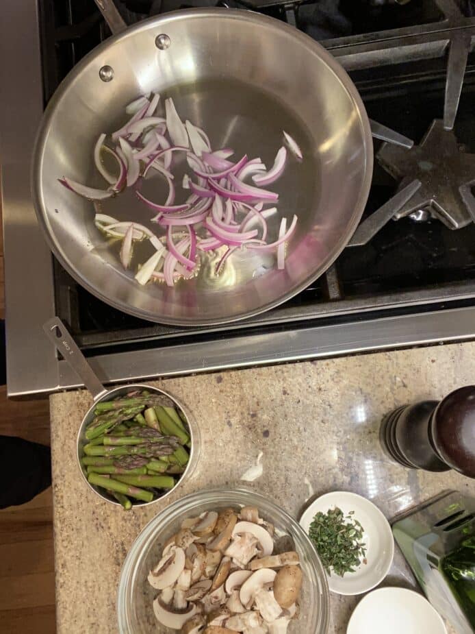 Red onions cooking in a large saute pan on the stove. The other ingredients for the asparagus mushroom saute are sitting on the side and ready to be added.