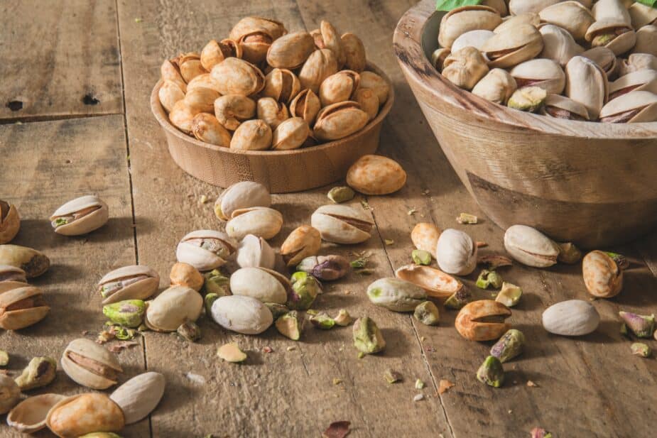 Pistachio nuts in the shell and out of the shell on a wooden table. Additional nuts with seasoning on top are in a small wooden bowl and more unseasoned in a large wooden bowl next to it.