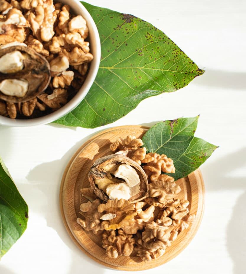 Walnuts and a walnut shell cracked open onin a small white bowl and on top of a small wooden board with walnut tree leaves. The plate and bowl are sitting on a white counter