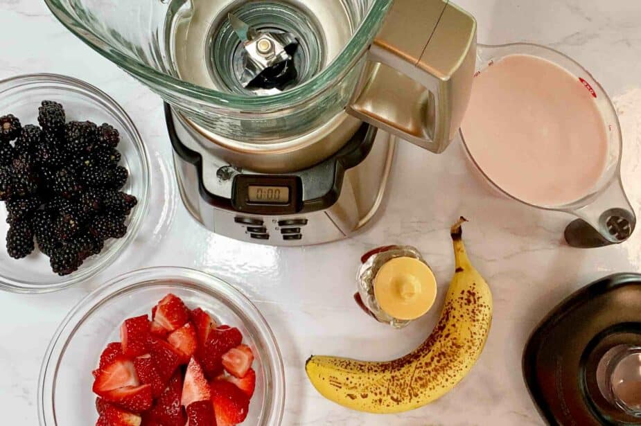 A glass bowl of strawberries, one with blackberries, a banana, measuring cup full of berry kefir and a blender on a marble counter.