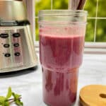 A bright pink smoothie in a glass smoothie cup with a metal straw. A blender in the background. Fresh blackberries and mint is on the counter in front of the smoothie.