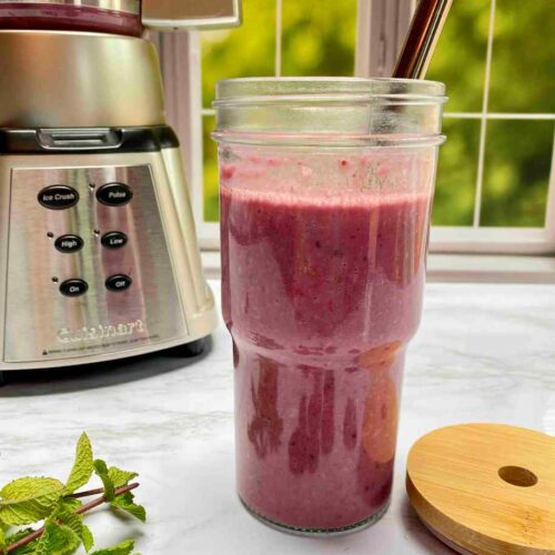 A bright pink smoothie in a glass smoothie cup with a metal straw. A blender in the background. Fresh blackberries and mint is on the counter in front of the smoothie.