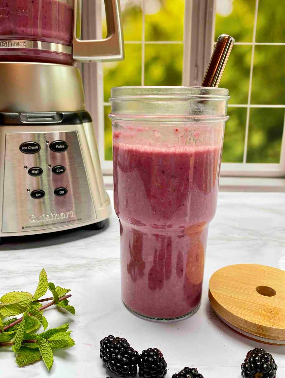 A bright pink smoothie in a glass smoothie cup with a metal straw. A blender in the background. Fresh blackberries and mint is on the counter in front of the smoothie.