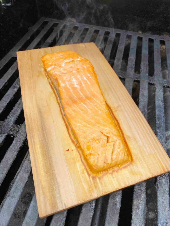 A fillet of salmon on a cedar plank sitting on gas grill grates with a little swirl of smoke rising from the salmon.