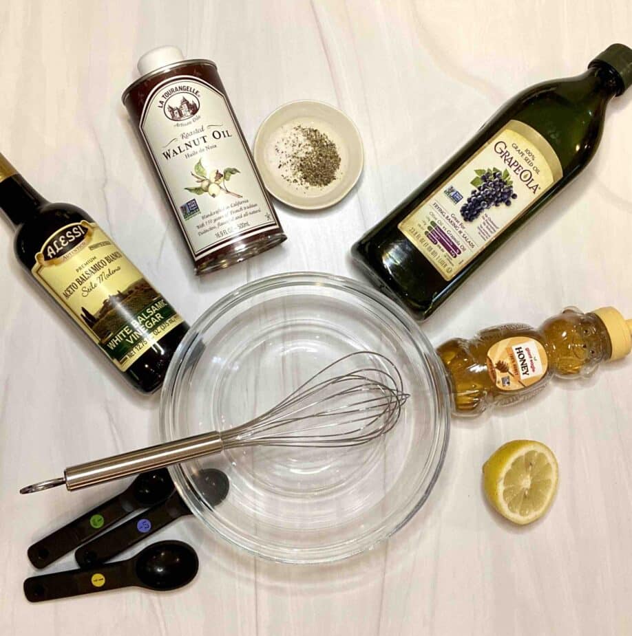 Walnut oil, balsamic vinegar, grapeseed oil, honey and a lemon on a marble counter with a mixing bowl and whisk in the middle.