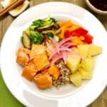 A white bowl filled with brown rice and quinoa with chunks of grilled salmon, pineapple pieces, bell pepper slices and sauteed greens and mushrooms.