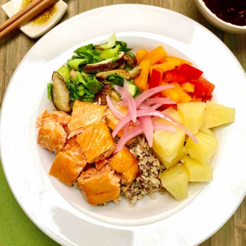 A white bowl filled with brown rice and quinoa with chunks of grilled salmon, pineapple pieces, bell pepper slices and sauteed greens and mushrooms.