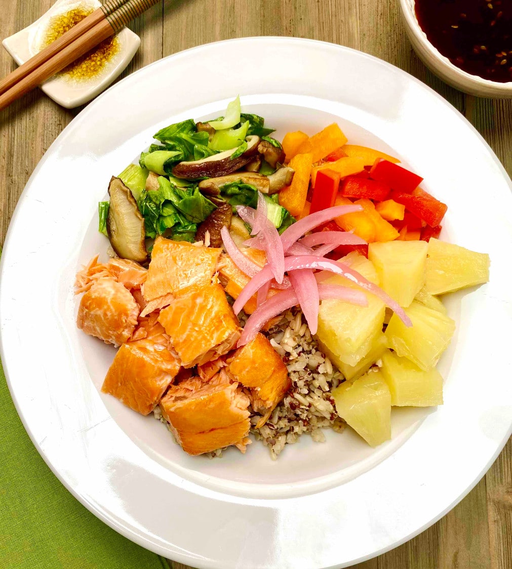 A white bowl filled with brown rice and quinoa with chunks of grilled salmon, pineapple pieces, bell pepper slices and sauteed greens and mushrooms.