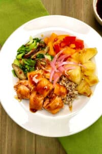 A white bowl filled with brown rice and quinoa with chunks of grilled salmon, pineapple pieces, bell pepper slices and sauteed greens and mushrooms.