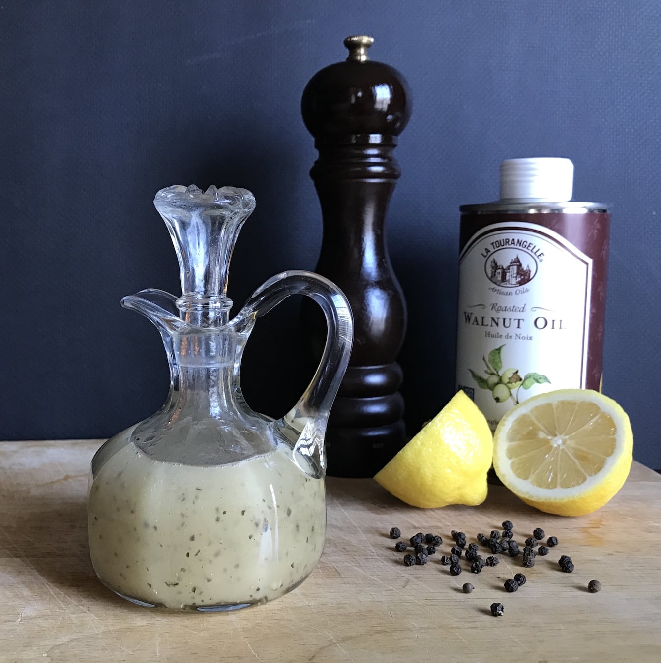 A glass carafe of walnut oil salad dressing with a pepper grinder, cut lemon, peppercorns and a bottle of walnut oil on a wood board.